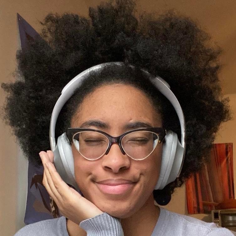 Aamina is smiling and cradles her chin in her hand. She has an afro and is wearing silver headphones.